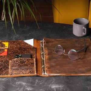 May include: Open leather journal with a brown floral embossed design, featuring a pen holder, card slots, and a phone pocket. Round eyeglasses rest on the open pages. A gray mug sits nearby. The journal is personalized with the name "Emily Thompson".