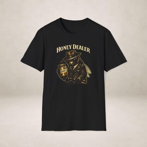 Op de afbeelding: Zwart t-shirt met een cartoonbij die een hoed en een pak draagt en een pot honing vasthoudt. De tekst "HONEY DEALER" staat boven de bij. Het ontwerp is in goud- en bruintinten.
