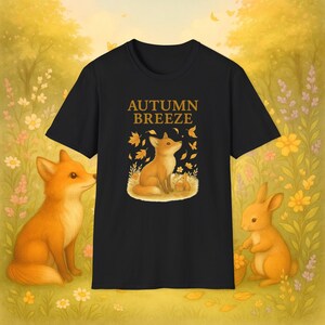 Op de afbeelding: Zwart t-shirt met de tekst "AUTUMN BREEZE" boven een afbeelding van een vos, bladeren en een klein dier. Twee oranje vossen en een konijn staan aan weerszijden van het shirt, tegen een gele en groene bloemenachtergrond.