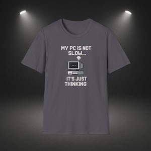 Op de afbeelding: Een grijs t-shirt met de tekst "MY PC IS NOT SLOW... IT'S JUST THINKING" en een pixelachtige computergrafiek. Het ontwerp toont een computermonitor, een CPU en een wolkengrafiek. Het shirt is een casual kledingstuk.