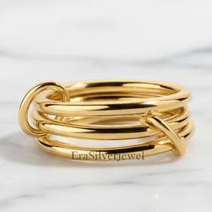 May include: A gold-colored ring with a multi-layered design. The ring features several connected bands, with a unique looped and crossed detail. The ring is displayed on a white marble surface. The text "EraSilverjewel" is visible.