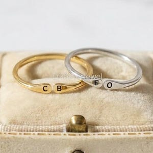 May include: Two delicate rings, one gold and one silver, rest on a beige velvet surface inside a small box. The gold ring has the letters "C B", and the silver ring has "F O". The rings are simple and elegant.