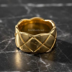 May include: A gold-colored ring with a quilted design. The ring has a wide band and a textured surface with a repeating diamond pattern. The ring is set against a dark, marbled background.