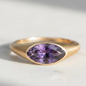 Op de afbeelding: Een gouden ring met een paarse marquise-geslepen edelsteen. De ring heeft een gladde, matte afwerking en een eenvoudig, elegant ontwerp. De edelsteen is gezet in een bezel setting.