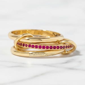 May include: A gold-colored ring set with three interlocking bands. One band is adorned with small, vibrant red gemstones. The ring is displayed on a white marble surface. The jewelry is elegant and eye-catching.