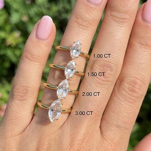 Pode incluir: Uma mão exibe quatro anéis de ouro com diamantes de corte marquesa de vários tamanhos: 1,00 CT, 1,50 CT, 2,00 CT e 3,00 CT. Os anéis têm bandas de ouro. O fundo apresenta folhas verdes e flores cor-de-rosa.