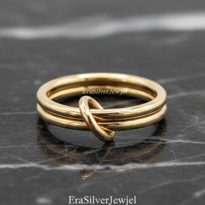 May include: A gold-colored ring with a knot design. The ring features two bands, with a knot detail at the top. The ring is displayed on a dark, marbled surface. The text "EraSilverJewjel" is visible on the ring.
