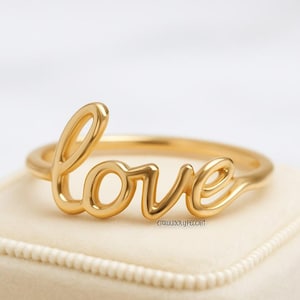 Può includere: Un anello dorato con la parola "love" scritta in corsivo. L'anello è presentato su un cofanetto beige. Questo delicato gioiello è perfetto come regalo o per l'uso quotidiano.