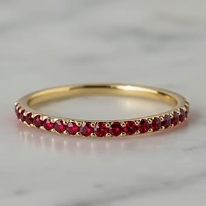 Könnte beinhalten: Ein goldener Bandring mit einer durchgehenden Reihe kleiner, runder, leuchtend roter Edelsteine. Der Ring ist vor einem verschwommenen, hellfarbigen Hintergrund platziert, der das Design und die Farbe des Rings hervorhebt.