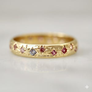 May include: A gold ring featuring a repeating pattern of stars and small, colourful gemstones. The gemstones are pink, purple, and peach-coloured. The ring has a smooth, rounded band.