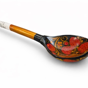 Può includere: Un cucchiaio di legno con un design floreale decorativo. La conca del cucchiaio è nera con motivi floreali rossi e gialli. Il manico è marrone chiaro con una punta rossa e un'etichetta bianca.