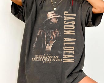 May include: Dark gray t-shirt featuring a graphic of Jason Aldean with a guitar and cowboy hat. The text "JASON ALDEAN" is printed vertically, and "HIGHWAY DESPERADO TOUR" is printed at the bottom. The shirt has a vintage, worn look.