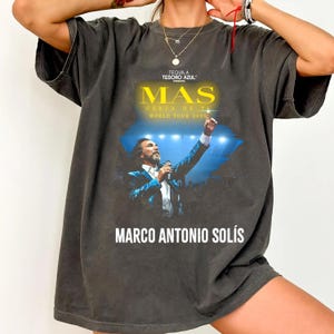 Peut inclure: T-shirt gris foncé avec un graphisme de concert. Le graphisme comprend le texte "MAS" en jaune, les mots "CERCA DE TI WORLD TOUR 2025", et le nom "MARCO ANTONIO SOLÍS". L'image montre un homme en train de chanter.
