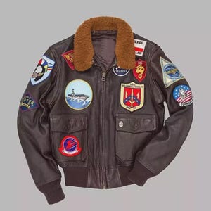 May include: A brown leather bomber jacket with a shearling collar and multiple embroidered patches. The jacket features a zip-up front, two front pockets, and ribbed cuffs and hem. Patches include military and naval insignia.