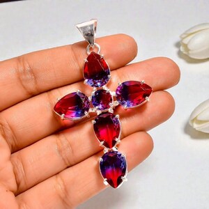 May include: A cross-shaped pendant featuring five pear-cut gemstones and one round-cut gemstone set in silver. The stones exhibit a gradient color transition from deep red to vibrant purple. A silver bail is attached to the top of the cross.