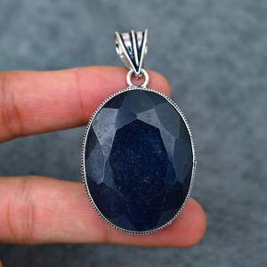 May include: Oval-shaped, faceted, dark blue gemstone pendant with a silver-coloured bail. The gemstone is set in a silver-coloured bezel with a dotted border. The pendant is held by a hand against a grey background. A piece of jewellery.