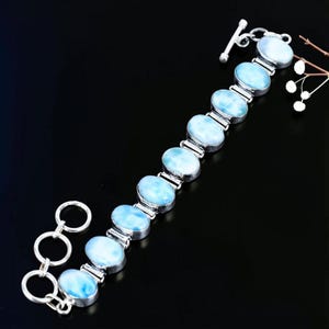 Könnte beinhalten: Ein Silberarmband mit neun ovalen, hellblau und weiß marmorierten Steinen in polierten Metallfassungen. Das Armband verfügt über einen Knebelverschluss, der aus einem runden Ring und einem T-förmigen Stab am anderen Ende besteht.