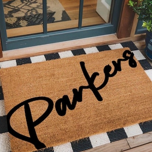 May include: A rectangular doormat with the name "Parkers" in black script. The mat is made of a natural brown material and has a black and white checkered border. The mat is placed in front of a door.