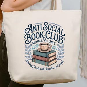 May include: Cream-colored tote bag featuring the text "Anti Social Book Club Member Only" in a whimsical font. The design includes a stack of books, a coffee cup, and the phrase "Making friends with characters, not people."
