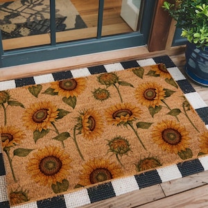 May include: A rectangular doormat with a sunflower pattern, set on top of a black and white striped mat. The sunflower mat has a tan background with yellow and brown sunflowers. The mats are placed in front of a door.