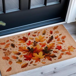 May include: Rectangular doormat with a tan background and a vibrant autumn leaf design. The leaves are in shades of red, orange, and yellow, creating a seasonal welcome. The mat is placed in front of a black door.