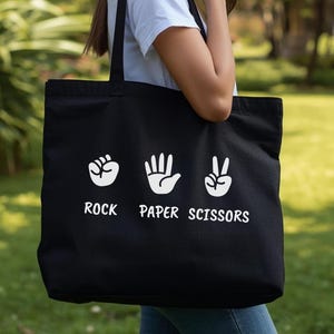 May include: Black tote bag with white graphic of rock, paper, scissors hand gestures and text. The bag has two black handles and is held by a person. The background is a green, blurred outdoor scene.