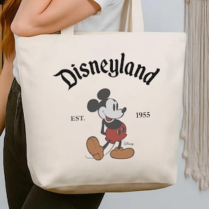 Op de afbeelding: Cr&egrave;mekleurige draagtas met het Disneyland-logo in zwart script, Mickey Mouse en de tekst "EST. 1955". De tas heeft een eenvoudig ontwerp met lange handvatten om over de schouder te dragen.