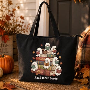 May include: A black tote bag featuring a graphic of several white ghosts sitting on a stack of colorful books, each reading a small book. The text "Read more books" is printed in white below the illustration. The bag has two black shoulder straps.