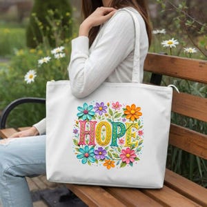 May include: White tote bag featuring the word "HOPE" in colorful, floral-style lettering. The word is surrounded by a vibrant array of flowers in shades of blue, pink, orange, and purple. The bag has a zipper closure and a shoulder strap.