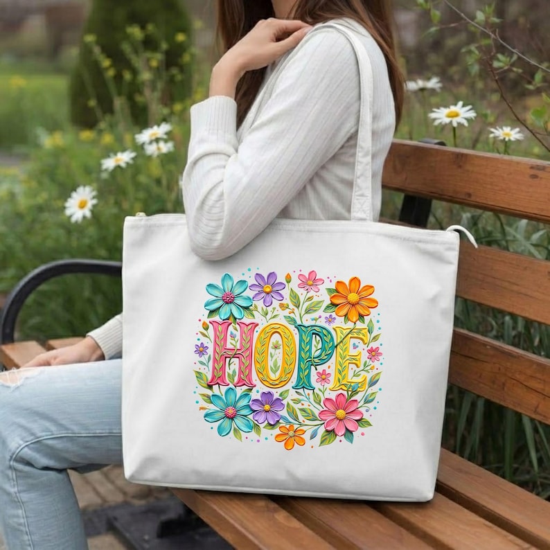 May include: White tote bag featuring the word "HOPE" in colorful, floral-style lettering. The word is surrounded by a vibrant array of flowers in shades of blue, pink, orange, and purple. The bag has a zipper closure and a shoulder strap.