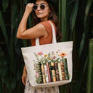 May include: Cream-colored tote bag featuring a floral and book design. The illustration includes a variety of colorful flowers and a row of vintage books. The bag has white handles and a zipper closure.