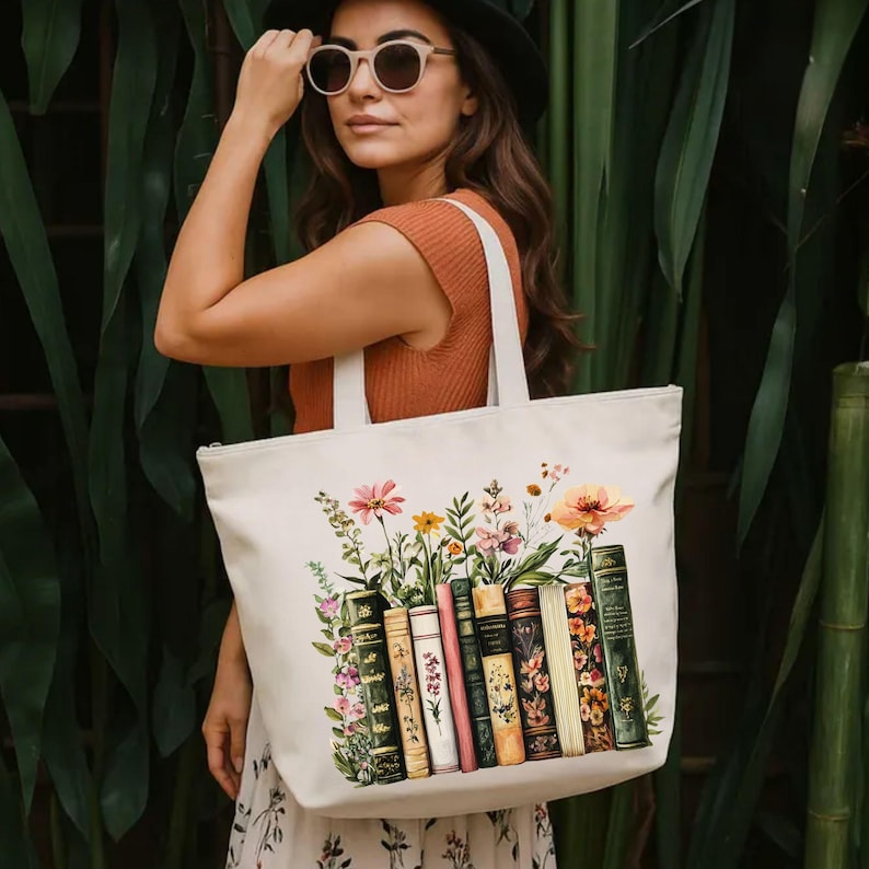 May include: Cream-colored tote bag featuring a floral and book design. The illustration includes a variety of colorful flowers and a row of vintage books. The bag has white handles and a zipper closure.