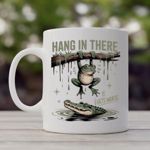May include: White ceramic mug featuring a cartoon frog hanging from a branch above an alligator in water. The mug has the text "HANG IN THERE" and "IT GETS WORSE". The design is in shades of green, brown, and beige.