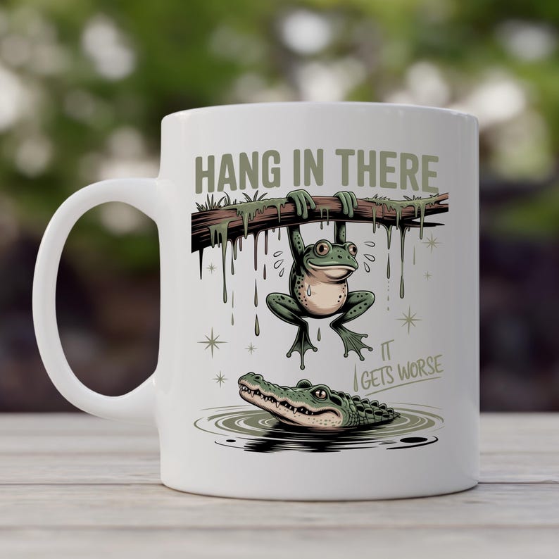 May include: White ceramic mug featuring a cartoon frog hanging from a branch above an alligator in water. The mug has the text "HANG IN THERE" and "IT GETS WORSE". The design is in shades of green, brown, and beige.