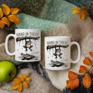 May include: Two white ceramic mugs with the phrase "HANG IN THERE" printed on them. Each mug features a cartoon frog hanging from a branch above an alligator in water. The phrase "IT GETS WORSE" is also printed on the mugs.
