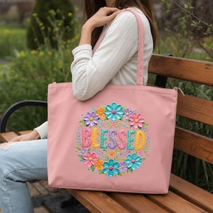 May include: A pink tote bag with a floral design and the word "BLESSED" in colorful letters. The flowers are in shades of pink, blue, orange, and purple. The bag has a zipper closure and pink straps.