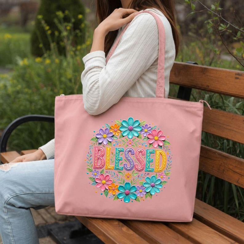 May include: A pink tote bag with a floral design and the word "BLESSED" in colorful letters. The flowers are in shades of pink, blue, orange, and purple. The bag has a zipper closure and pink straps.