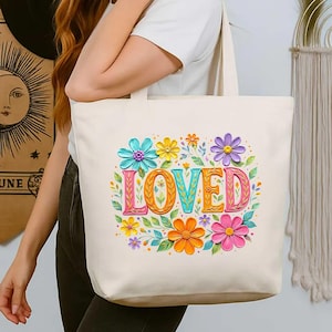 May include: Cream-colored tote bag featuring the word "LOVED" in colorful, textured letters. The word is surrounded by vibrant flowers in shades of blue, yellow, orange, pink, and purple, with green leaves. The bag has two handles.