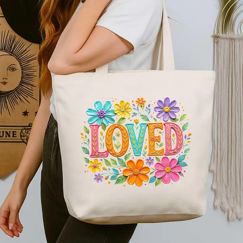 May include: Cream-colored tote bag featuring the word "LOVED" in colorful, textured letters. The word is surrounded by vibrant flowers in shades of blue, yellow, orange, pink, and purple, with green leaves. The bag has two handles.