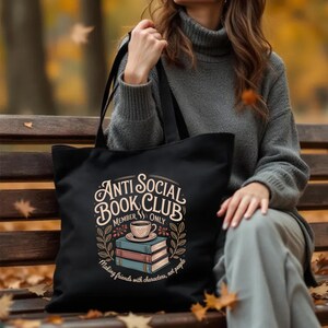 May include: Black tote bag with the text "Anti Social Book Club Member Only" in a vintage style. The design includes a coffee cup, books, and floral elements. The bag is carried by a person wearing a gray sweater.