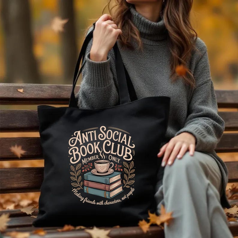May include: Black tote bag with the text "Anti Social Book Club Member Only" in a vintage style. The design includes a coffee cup, books, and floral elements. The bag is carried by a person wearing a gray sweater.