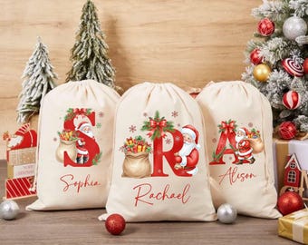 May include: Three cream-colored Christmas gift bags with red letter designs and Santa Claus illustrations. Each bag features a different name in red script: Sophia, Rachael, and Alison. The bags are decorated with festive bows and snowflakes.
