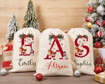 May include: Three cream-colored Christmas gift bags with red letters, Santa Claus illustrations, and names. The bags feature a festive design with snowflakes and greenery. The names "Emily", "Alison", and "Sophia" are written in cursive.