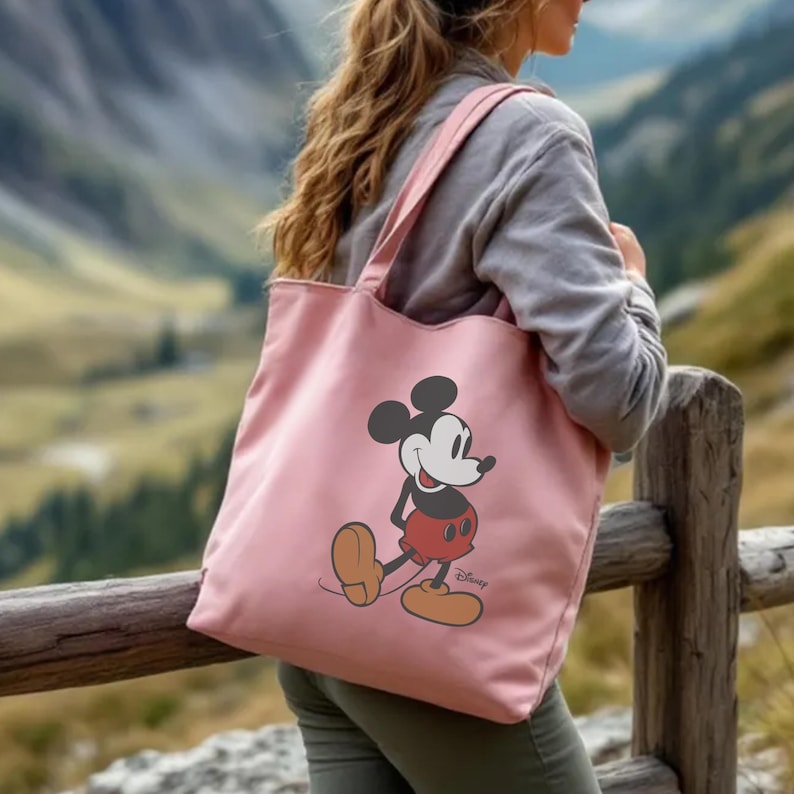 Puede incluir: Un bolso tote rosa con un dise&ntilde;o de Mickey Mouse. El bolso tiene correas largas para llevarlo al hombro. El personaje de dibujos animados est&aacute; representado con sus pantalones cortos rojos, cuerpo negro y zapatos amarillos. El bolso es de material suave.