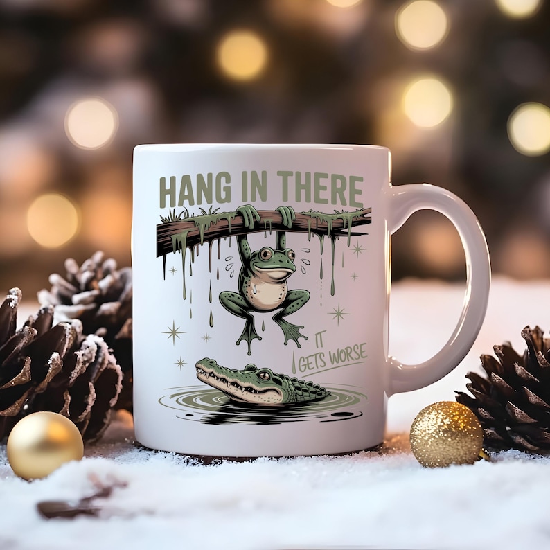May include: White ceramic mug with a humorous illustration of a frog hanging from a branch above an alligator in water. The mug has the text "HANG IN THERE" and "IT GETS WORSE" in green.