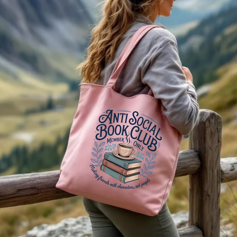 May include: A pink tote bag with the text "Anti Social Book Club Member Only" and a graphic of books and a coffee cup. The bag also has the text "Making friends with characters, not people". The bag is being carried outdoors.
