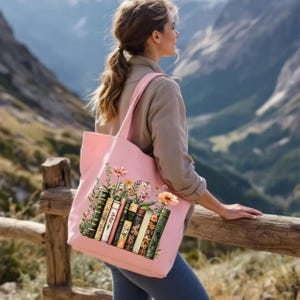 May include: A light pink tote bag featuring a design of vintage books and flowers. The bag has a rectangular shape with two shoulder straps. The books are in various colors and styles, with floral accents.
