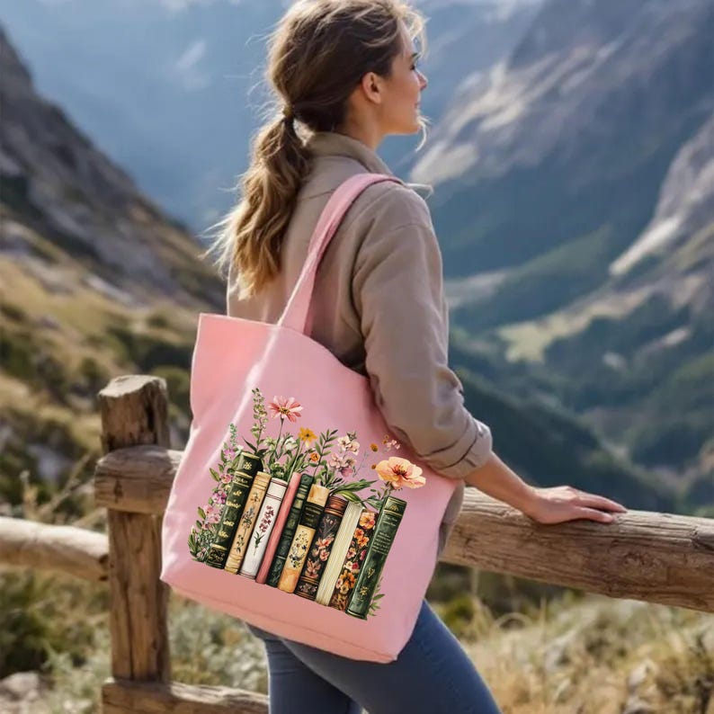May include: A light pink tote bag featuring a design of vintage books and flowers. The bag has a rectangular shape with two shoulder straps. The books are in various colors and styles, with floral accents.