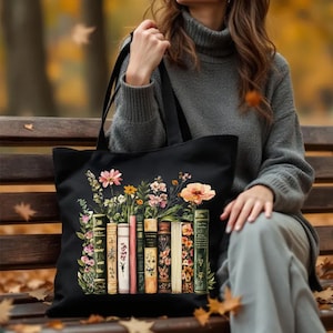 May include: Black tote bag featuring a floral design with a row of vintage books. The flowers include pink, yellow, and orange blooms. The books have various colors and floral patterns. The bag has black handles and is held by a person wearing a gray sweater.