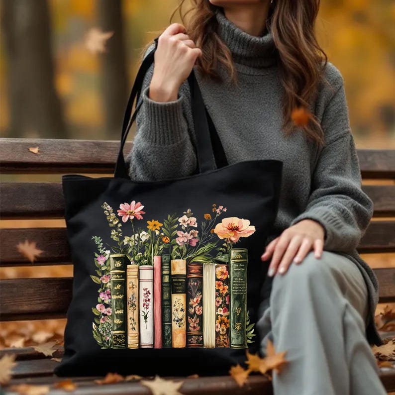 May include: Black tote bag featuring a floral design with a row of vintage books. The flowers include pink, yellow, and orange blooms. The books have various colors and floral patterns. The bag has black handles and is held by a person wearing a gray sweater.